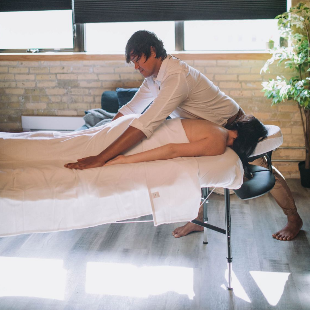 A massage therapist is placing their hands on a client who is laying face down draped with a white cotton sheet on a massage plinth 